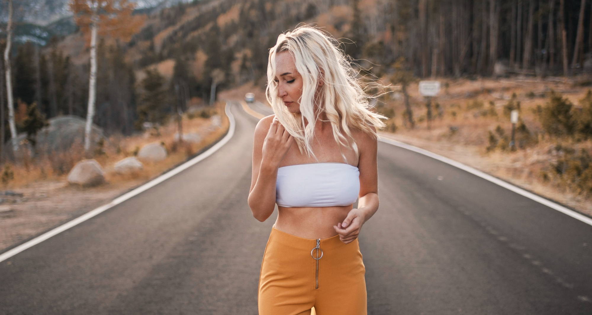  A woman with blond hair wearing orange pants and a white strapless top walks along a paved road in the mountains.