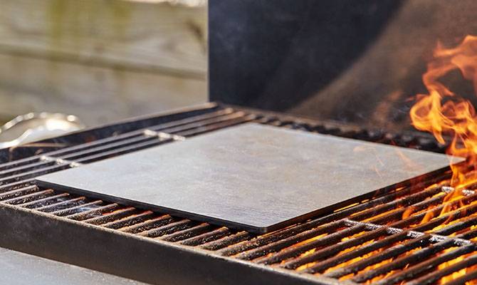 An empty Misen Oven Steel sits on an outdoor grill, with open flame visible in the foreground.