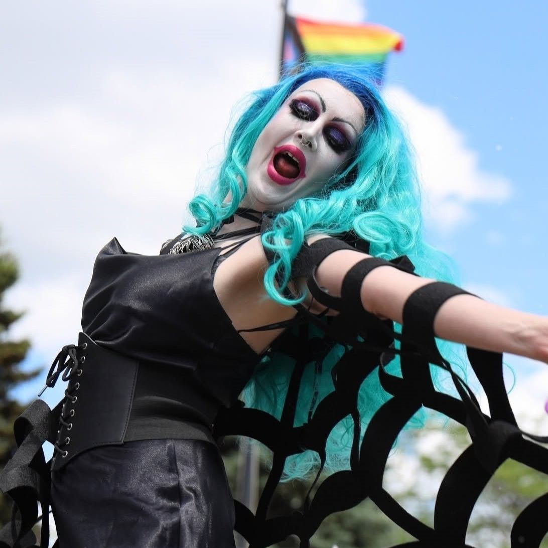 Person with blue hair in costume performing outdoors, with a rainbow flag in background.