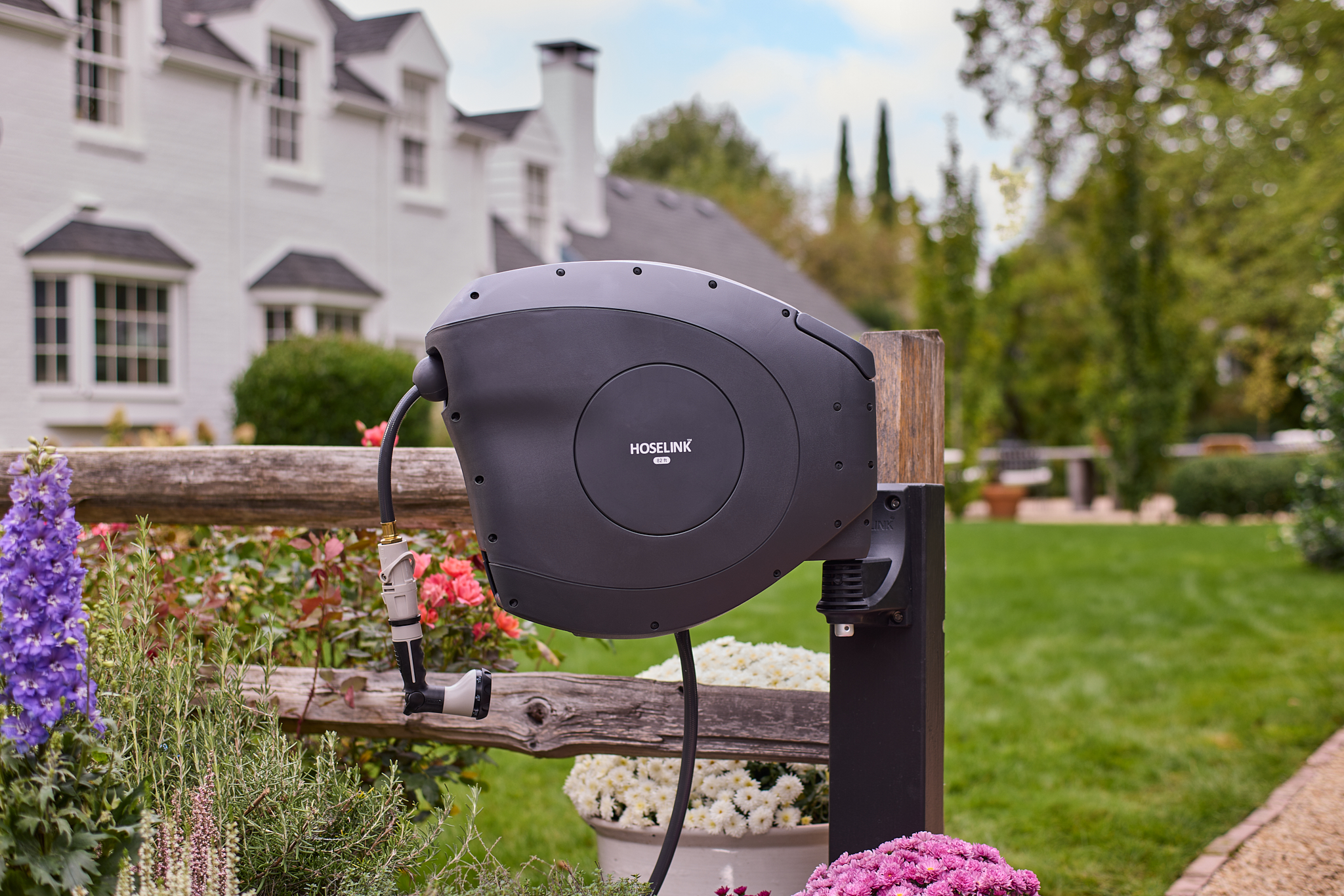 A charcoal colored Hoselink Retractable Hose Reel mounted on a wooden post near flowers.