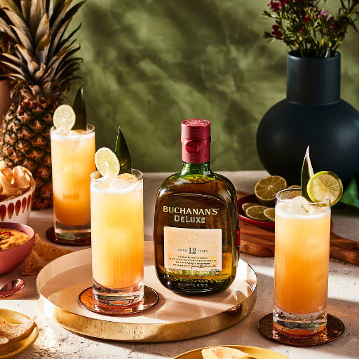 Three cocktails with a bottle of Buchanan's Deluxe and tropical decorations.