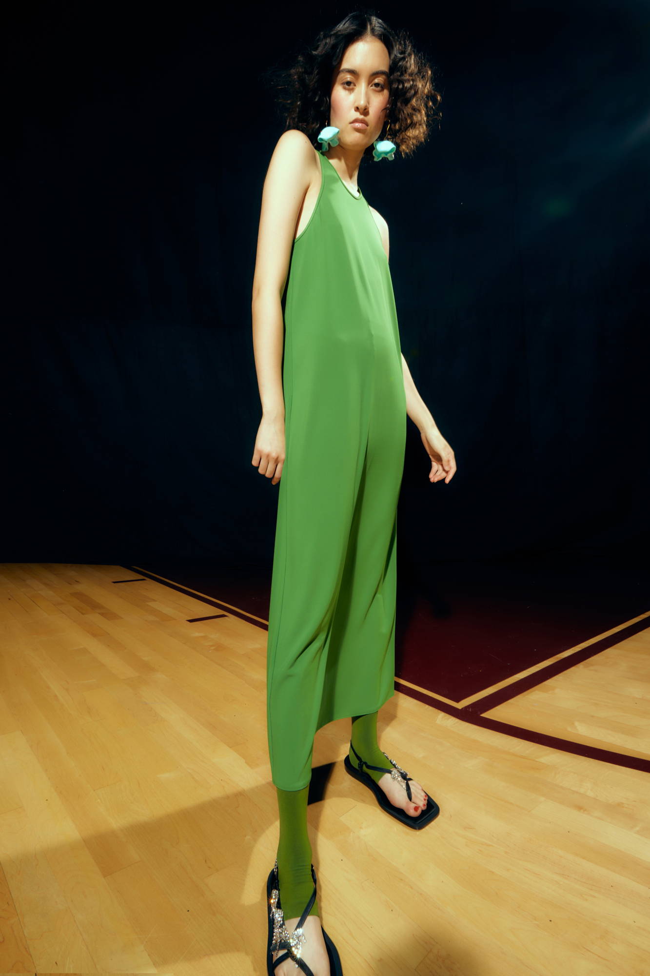 Model wearing a green dress and black sandals on a wooden floor.