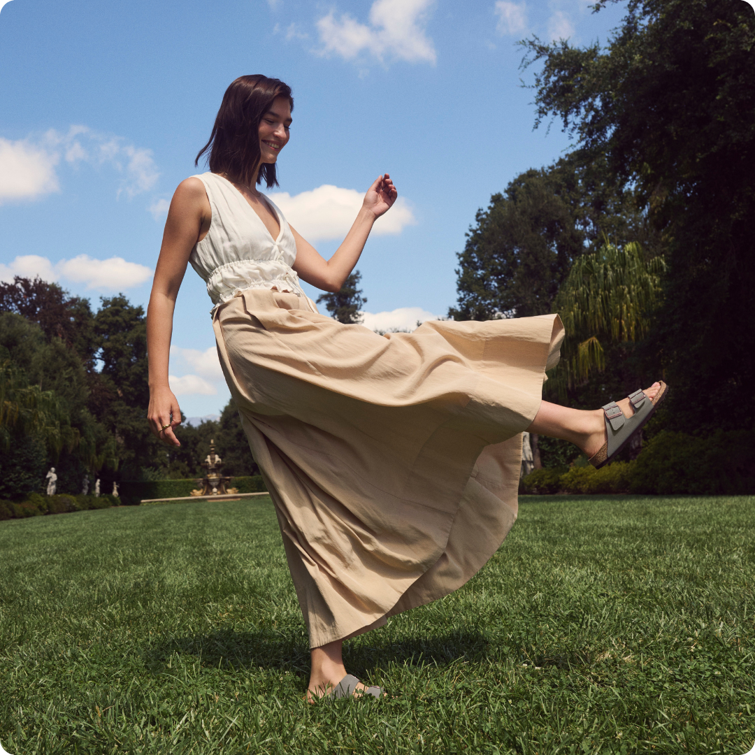 A woman in a long dress playfully walks on grass under a blue sky.