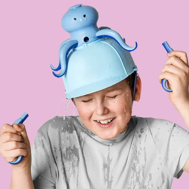 Child wearing a blue octopus water hat playing with water crayons, set against a pink background.