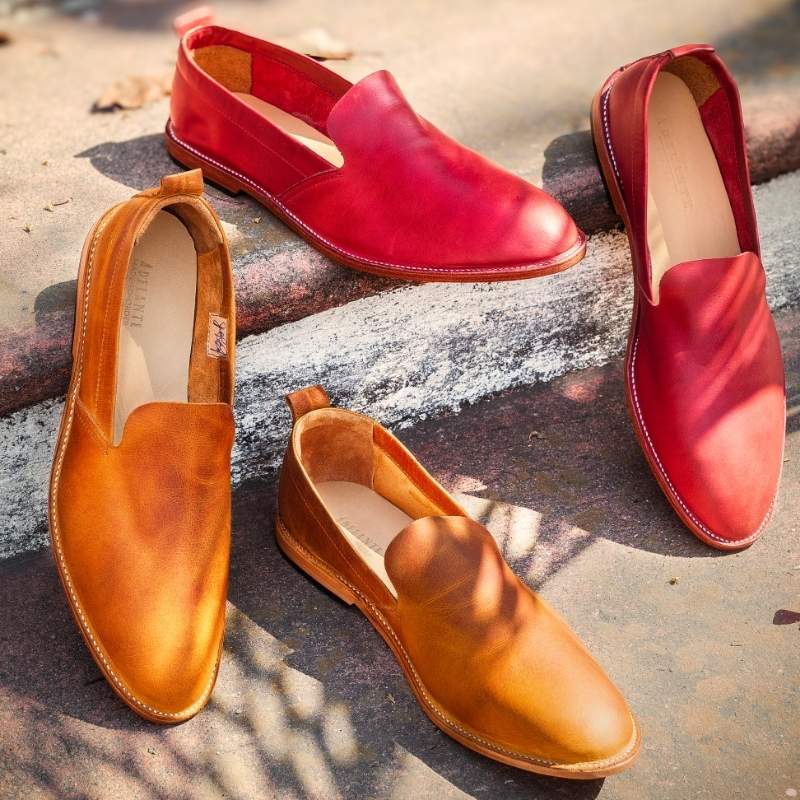 A top-down overhead shot of four pairs of handmade leather smoking loafers arranged neatly on rustic stone stairs under soft sunlight. The collection highlights color variations, showcasing two pairs of vibrant red leather loafers on the upper steps and two pairs of warm tan or light brown leather loafers on the lower steps.