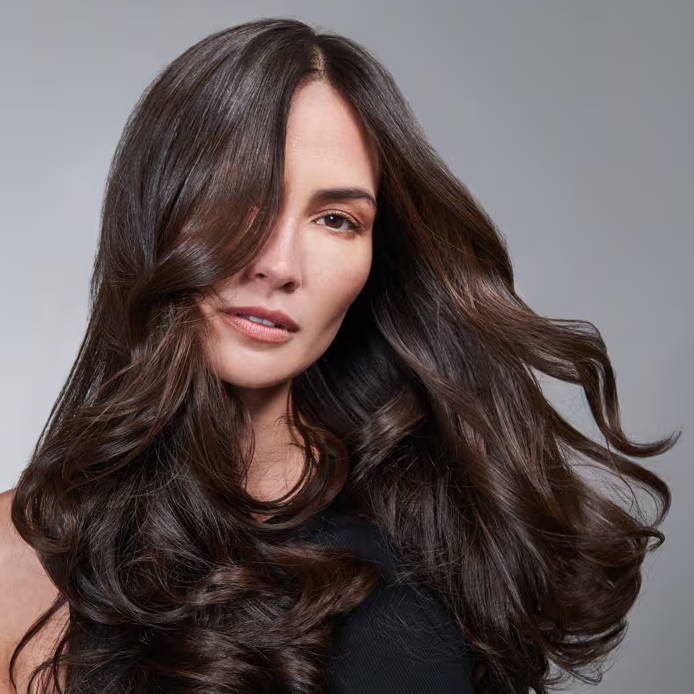 Woman with voluminous, wavy dark brown hair styled over one eye, posing against a gray background.
