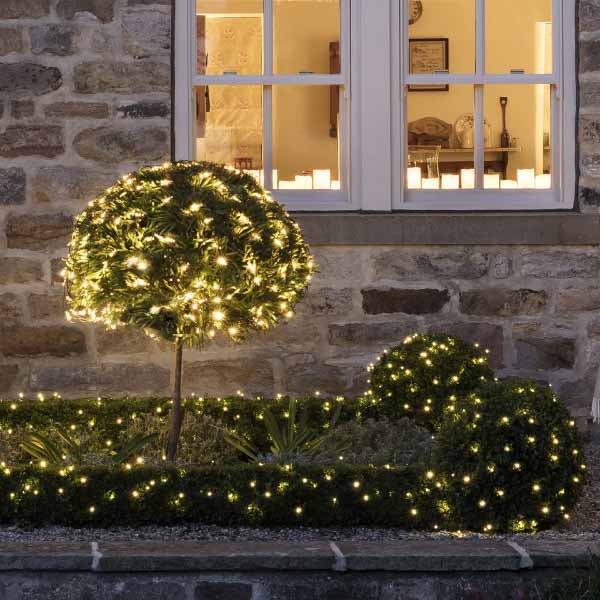 Outdoor plants and shrubs wrapped in lights outside a home.