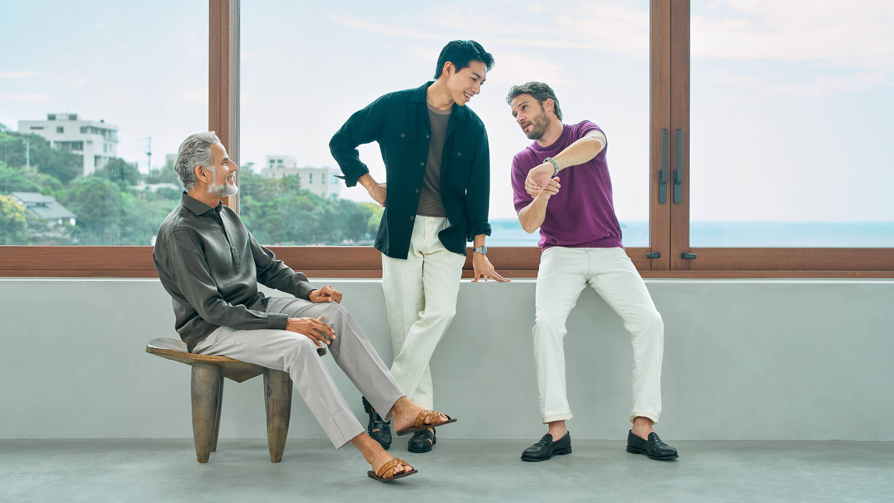 Three men in stylish outfits interacting indoors with a scenic view.