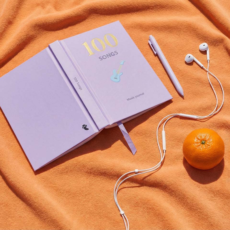 A lavender notebook, pen, earphones, and an orange on an orange blanket.