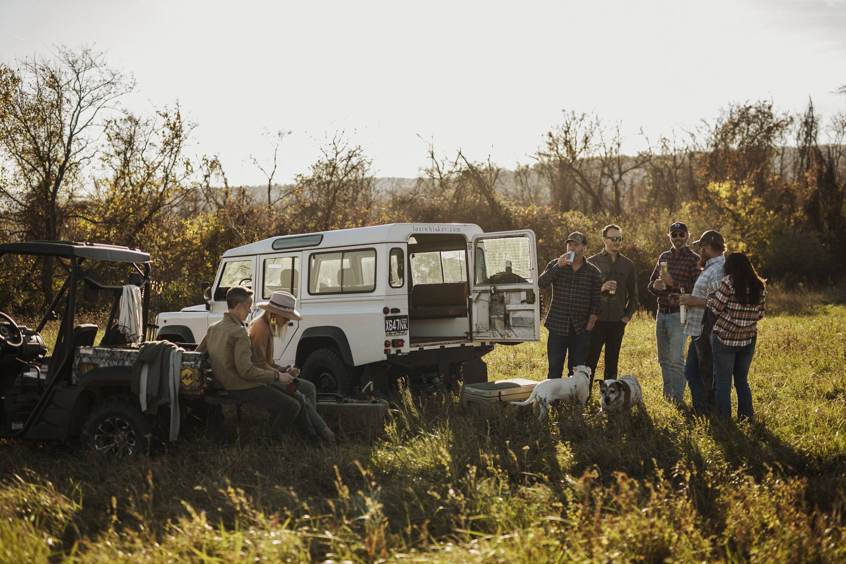 A group of people outdoors next to an antique vehicle and 4wd vehible drinking beer and having a good time.