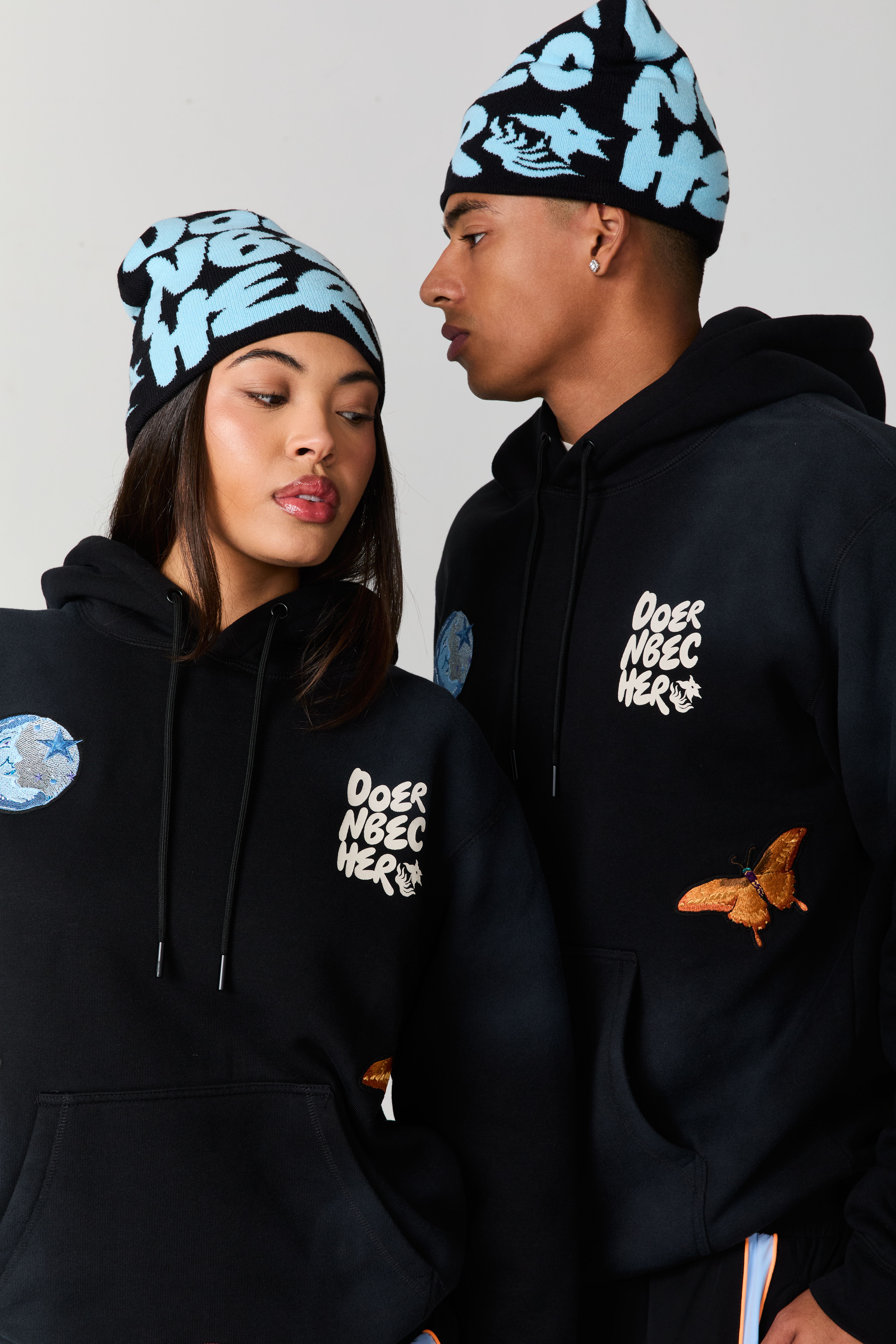 Two models wearing stylish hoodies and beanies, posing side by side.