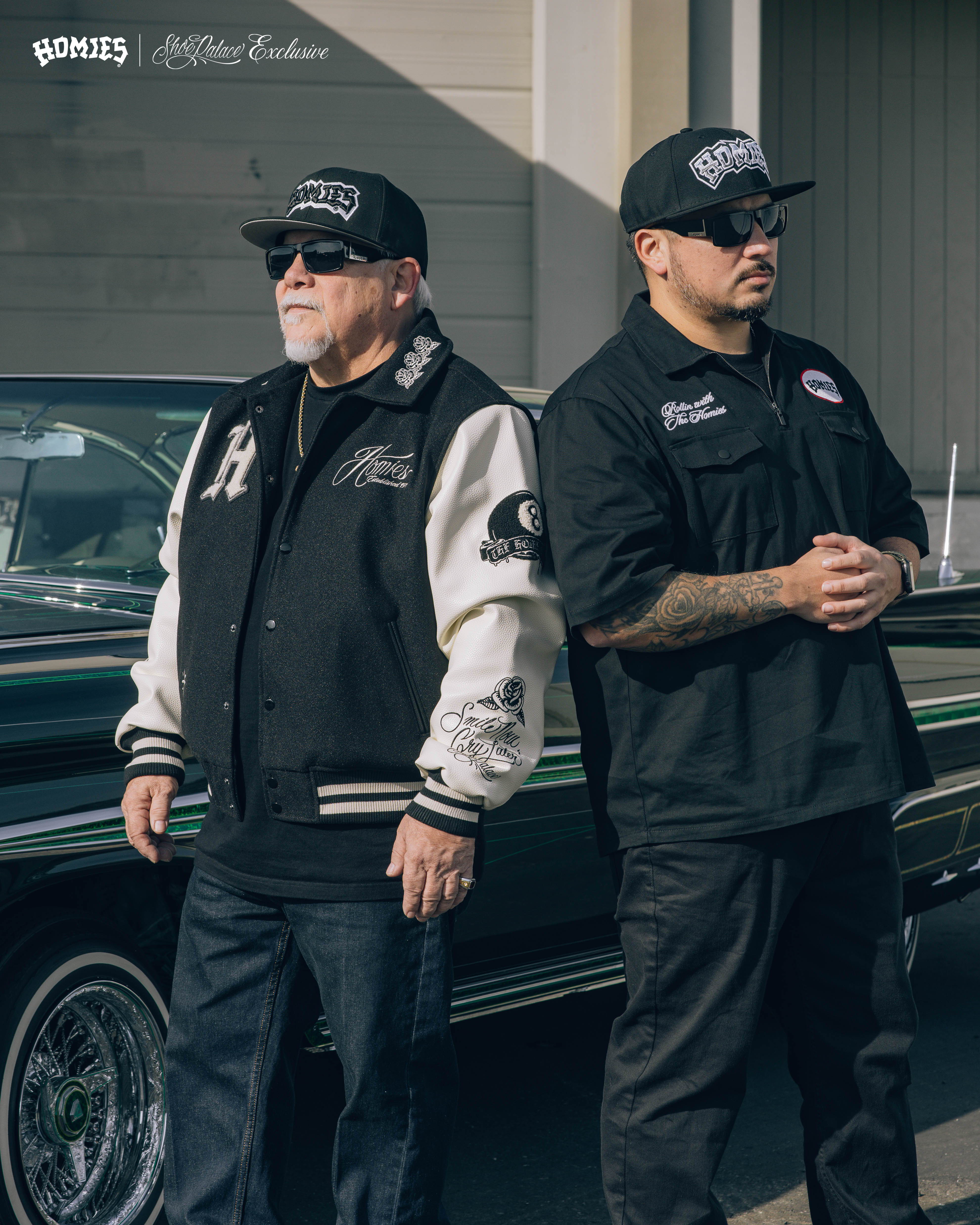 Two men standing beside a classic car, wearing matching jackets and hats.