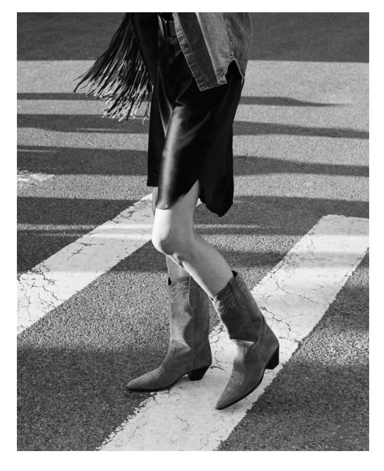 A person wearing cowboy boots crossing a street in black and white.
