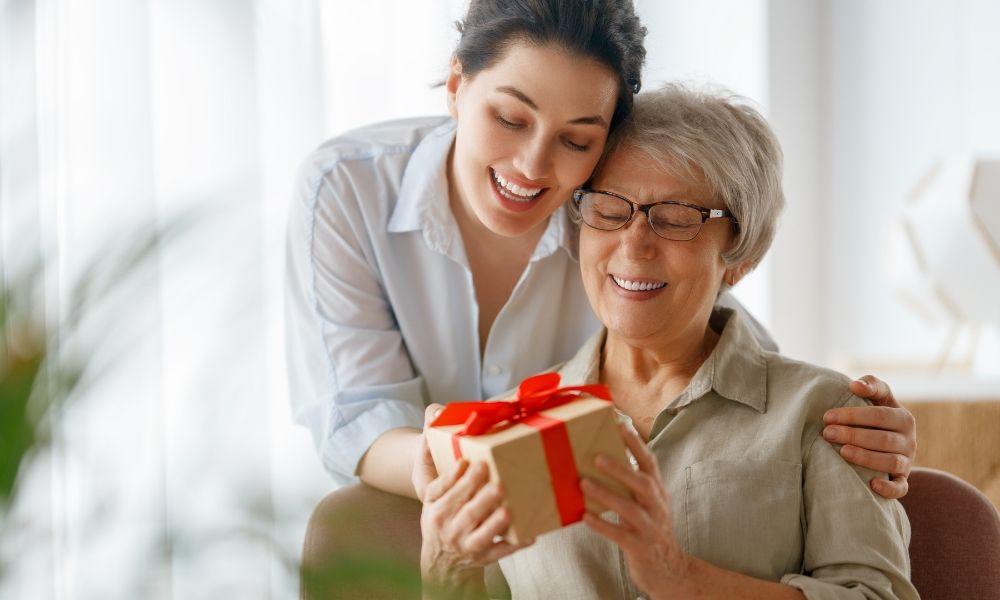 A woman gifts a present to an elderly lady, both smiling happily.