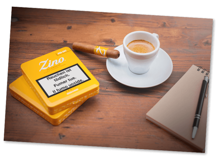 Eine Zino Nicaragua Half Corona-Zigarre mit innovativem Pre-Cut, die auf der Untertasse einer gefüllten Espressotasse liegt. Nebendran liegen ein Schreibblock mit Kugelschreiber und zwei Half Corona-Dosen.