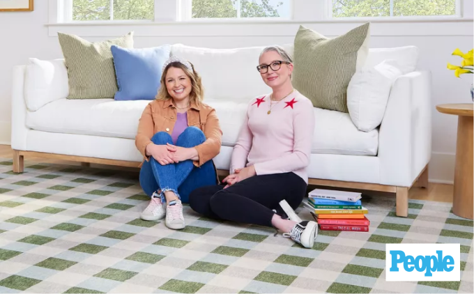 Joanna and Clea from The Home Edit sitting on a couch, surrounded by books on the floor.