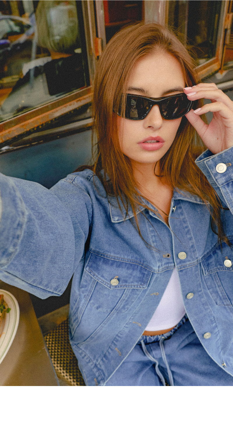 Young woman in denim jacket wearing sunglasses, taking a selfie.