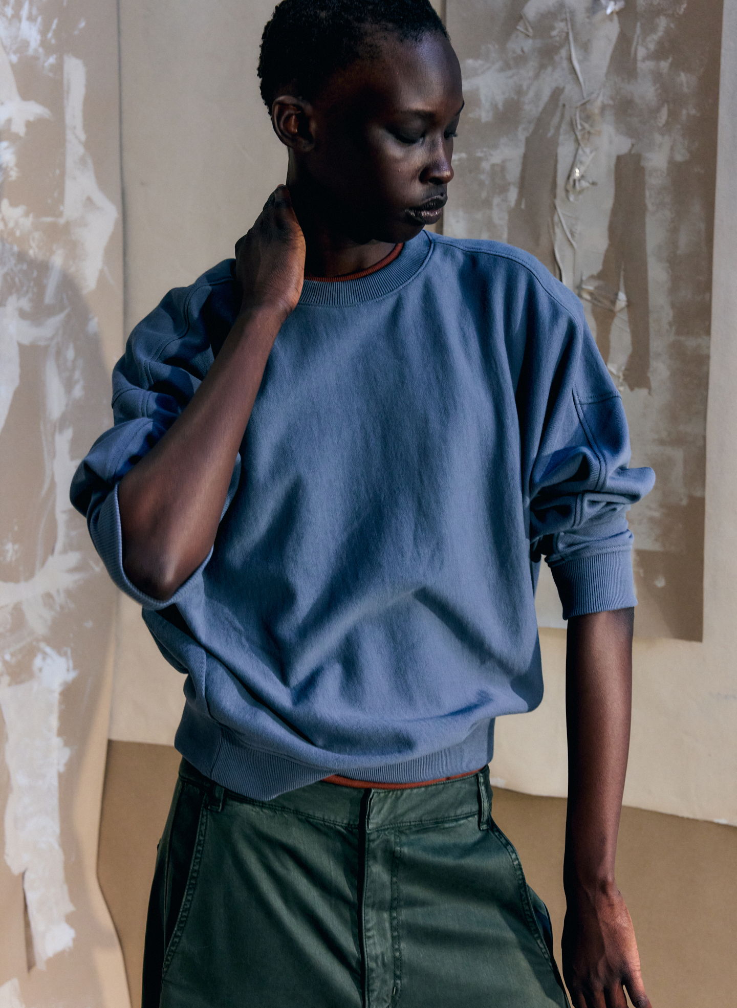 A person wearing a blue sweatshirt and green skirt, posing thoughtfully.