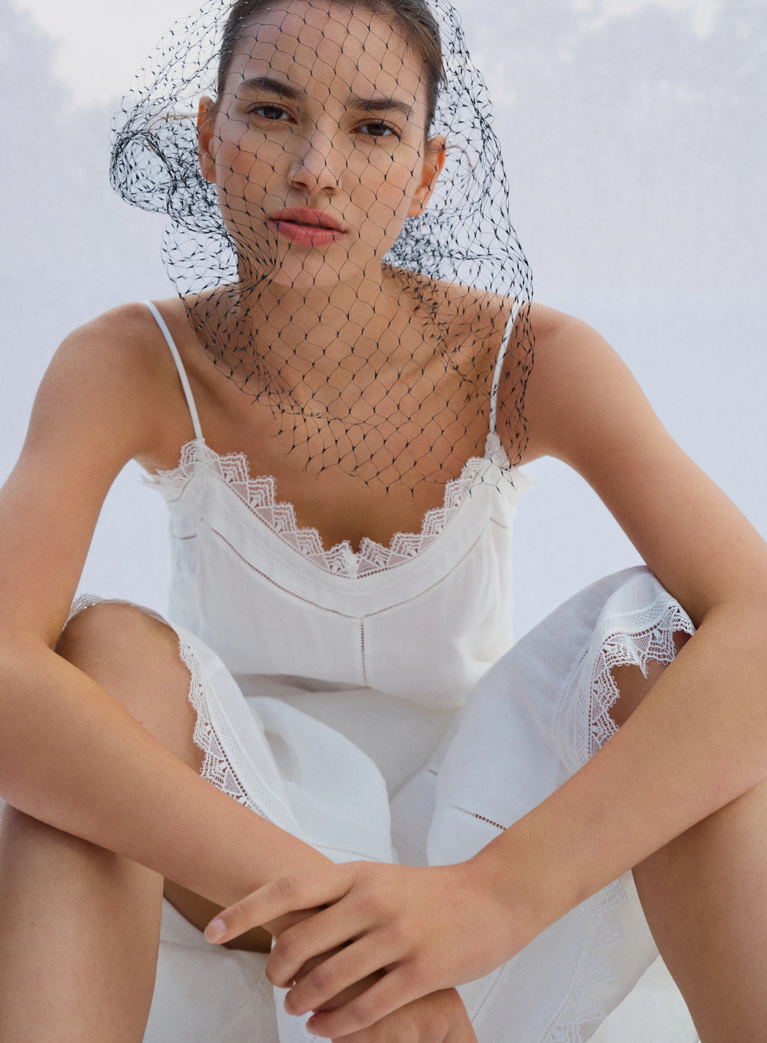 Model in white attire with a netted veil, posing thoughtfully.