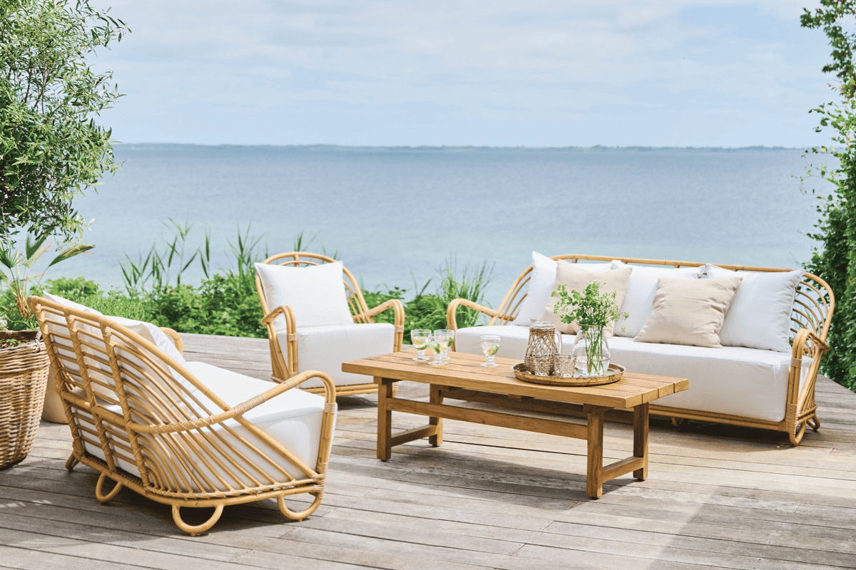 Outdoor wicker lounge chairs and sofa with white cushions arranged on a waterfront deck with a teak coffee table. 