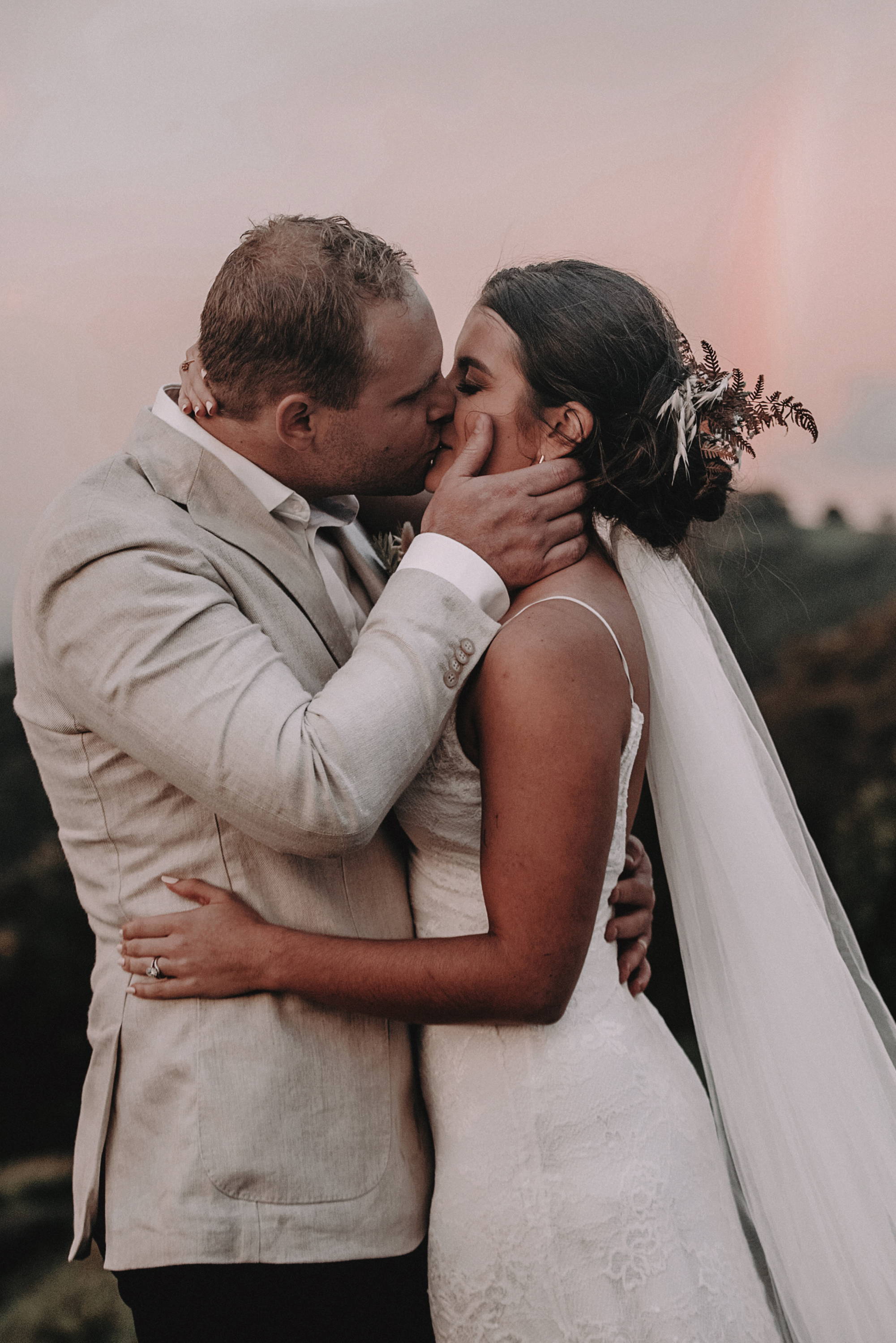Groom in a sand jacket kissing his bride wearing a thin strap lace dress