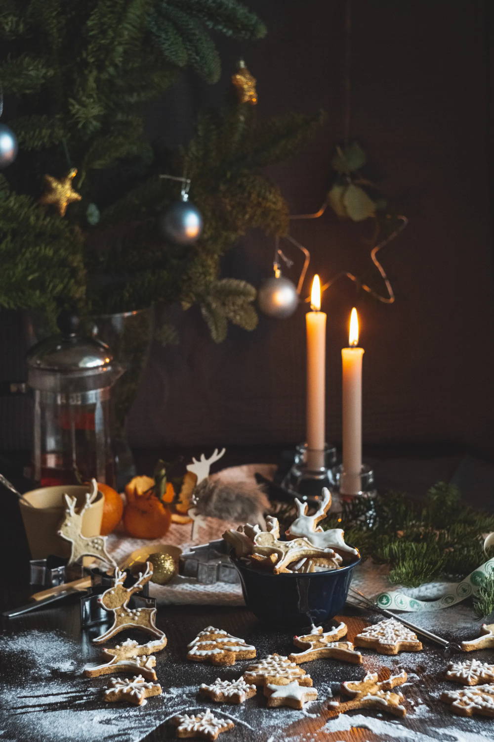 A counter dusted with flour and scattered with gingerbread cookies. In the background, two lit taper candles and several evergreen boughs.