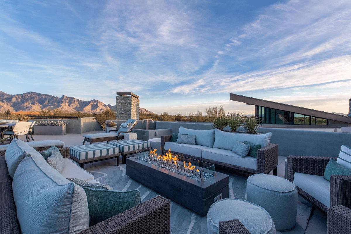 Rooftop outdoor living area with woven sofas around a rectangular fire table, with mountain views under a wide blue sky.