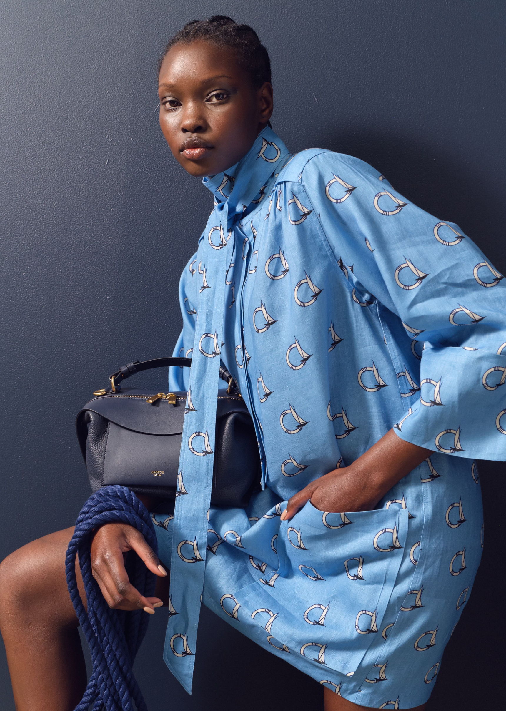 Model in a blue patterned dress, posing with a handbag against a dark background.