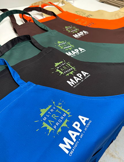 Stack of aprons in multiple colours including green, navy, black, and blue, each printed with the MAPA logo in white