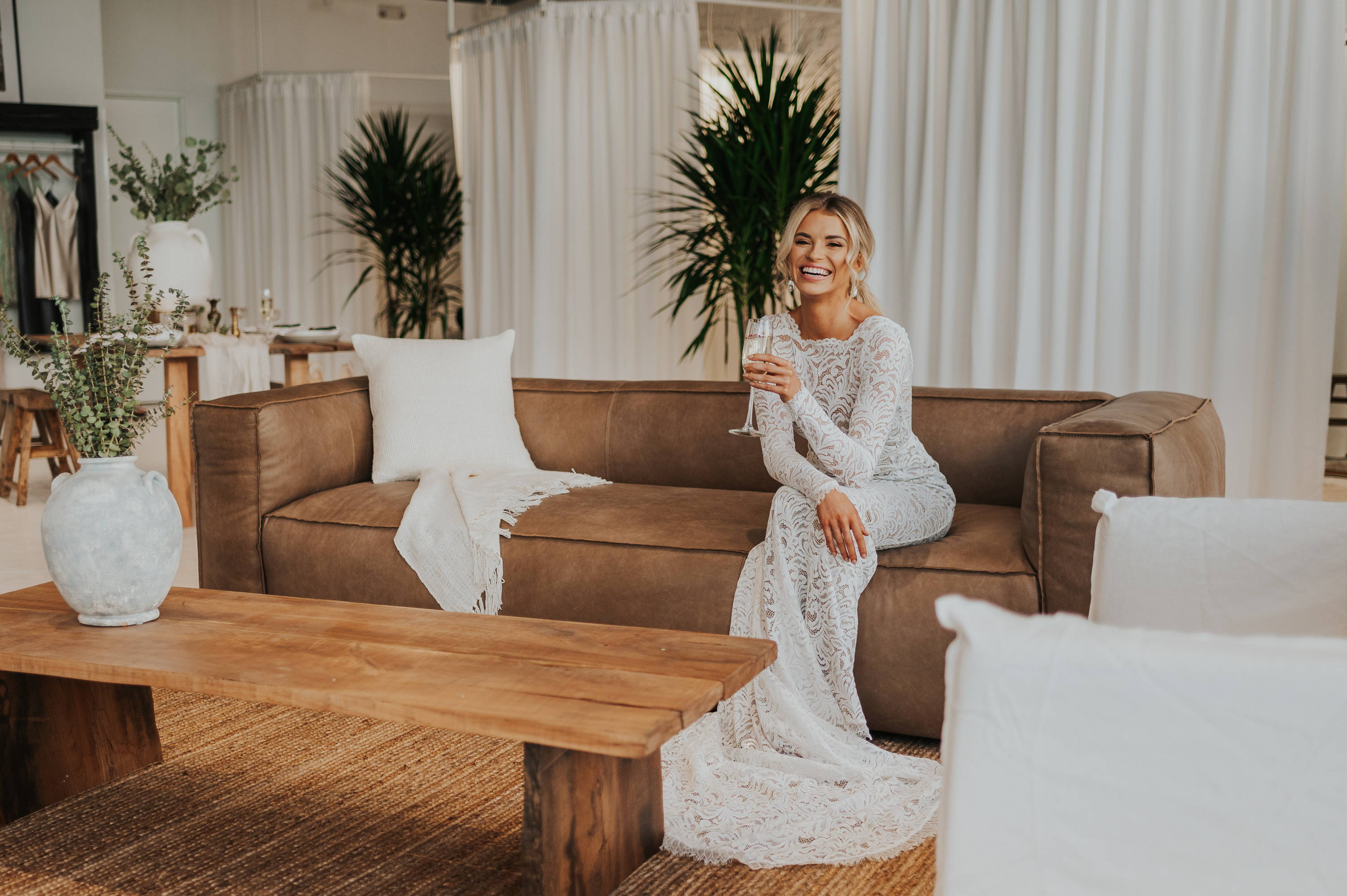 Die Braut trägt das Grace Loves Lace Orla Hochzeitskleid auf einer langen braunen Ledercouch mit einem Tisch aus recyceltem Holz