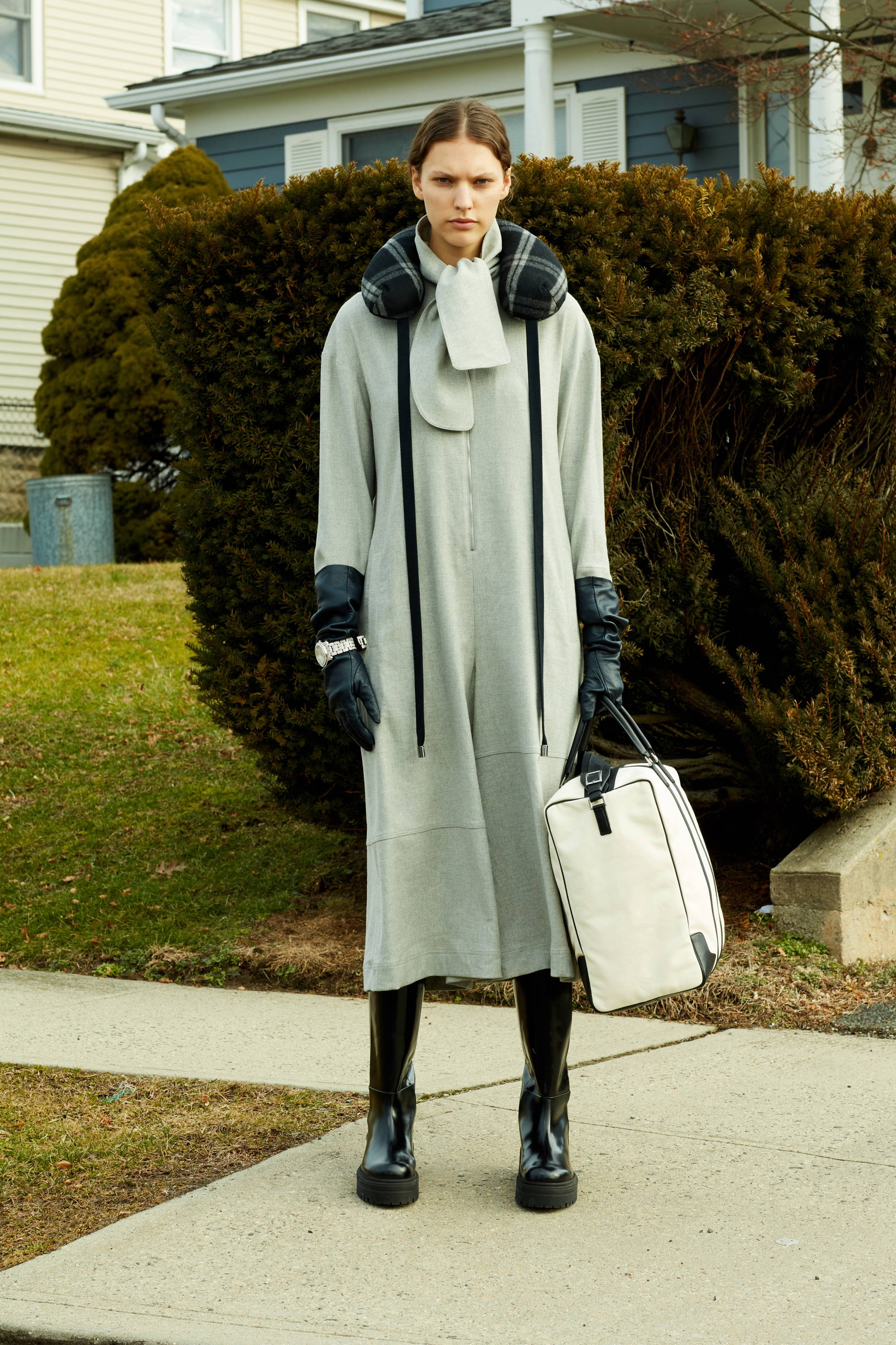 Model in a long gray coat, black gloves, and boots, holding a bag.