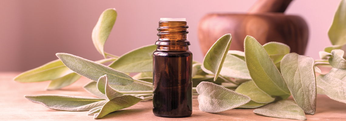 Brown essential oil bottle surrounded by green leaves on a wooden surface.