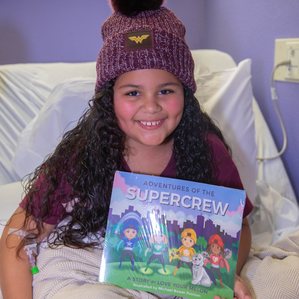 Young girl in a hospital bed wearing a Love Your Melon beanie smiling at the camera while holding up a children's book