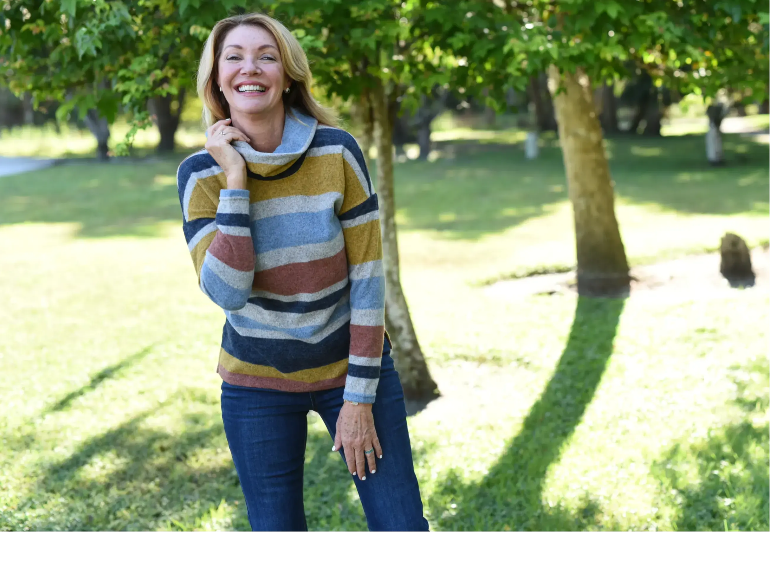 A smiling blonde woman in a striped sweater stands amongst some trees.