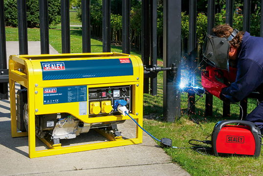 Welder using a generator to weld metal near a fence.