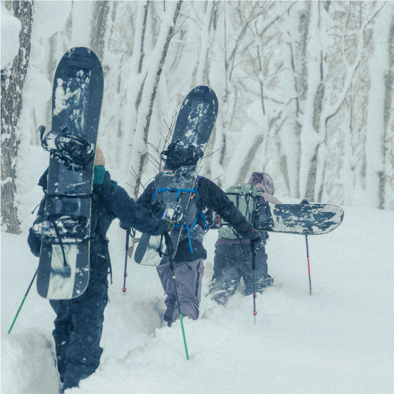 Four individuals hiking through deep snow, carrying snowboards in a snowy forest.