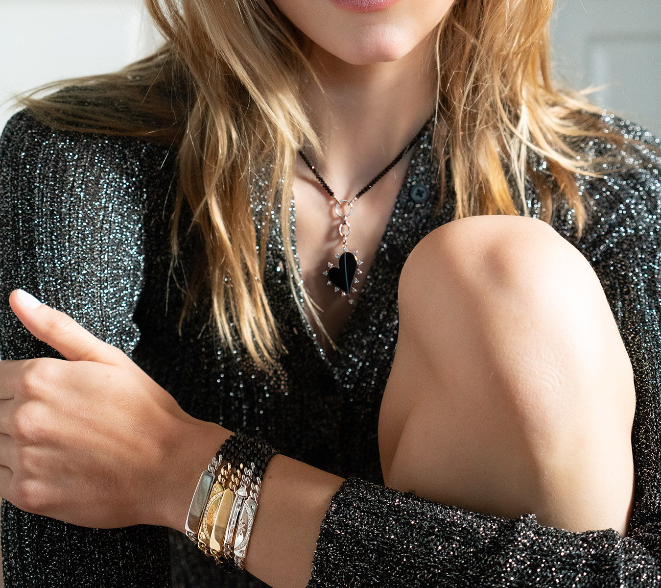 Woman wearing a black necklace and stacked bracelets, sitting with her knee raised.