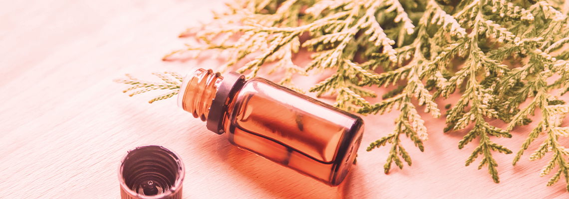 A bottle of essential oil next to green leaves on a pink background.