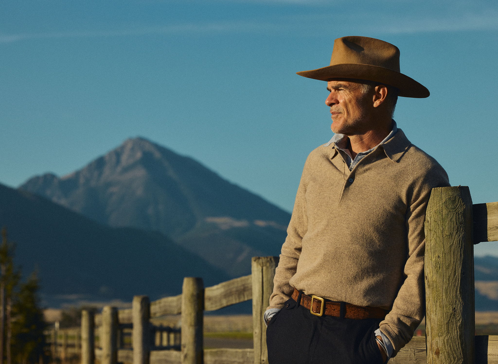 Michael Kelly leaning on a fence in Pray, Montana wearing a Vintage Stetson Hat, the Rivay Luca Italian Wool Sweater Polo in Camel over the Bridges Oxford Workshirt in Navy, a vintage leather belt and the Rivay Italian Wool Suit in Navy Chalk Stripe Trousers