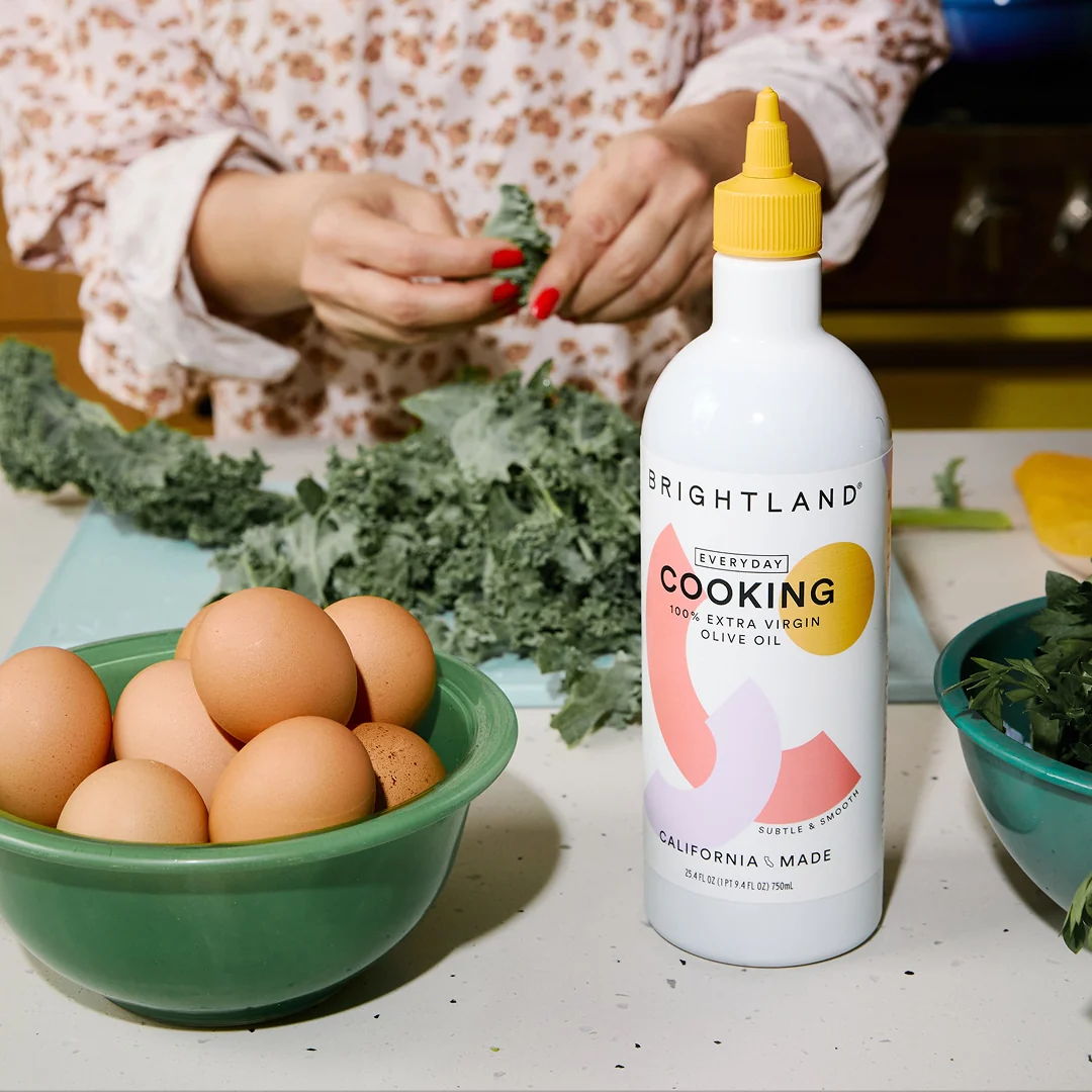 Person preparing food with eggs, greens, and cooking oil on a kitchen counter.