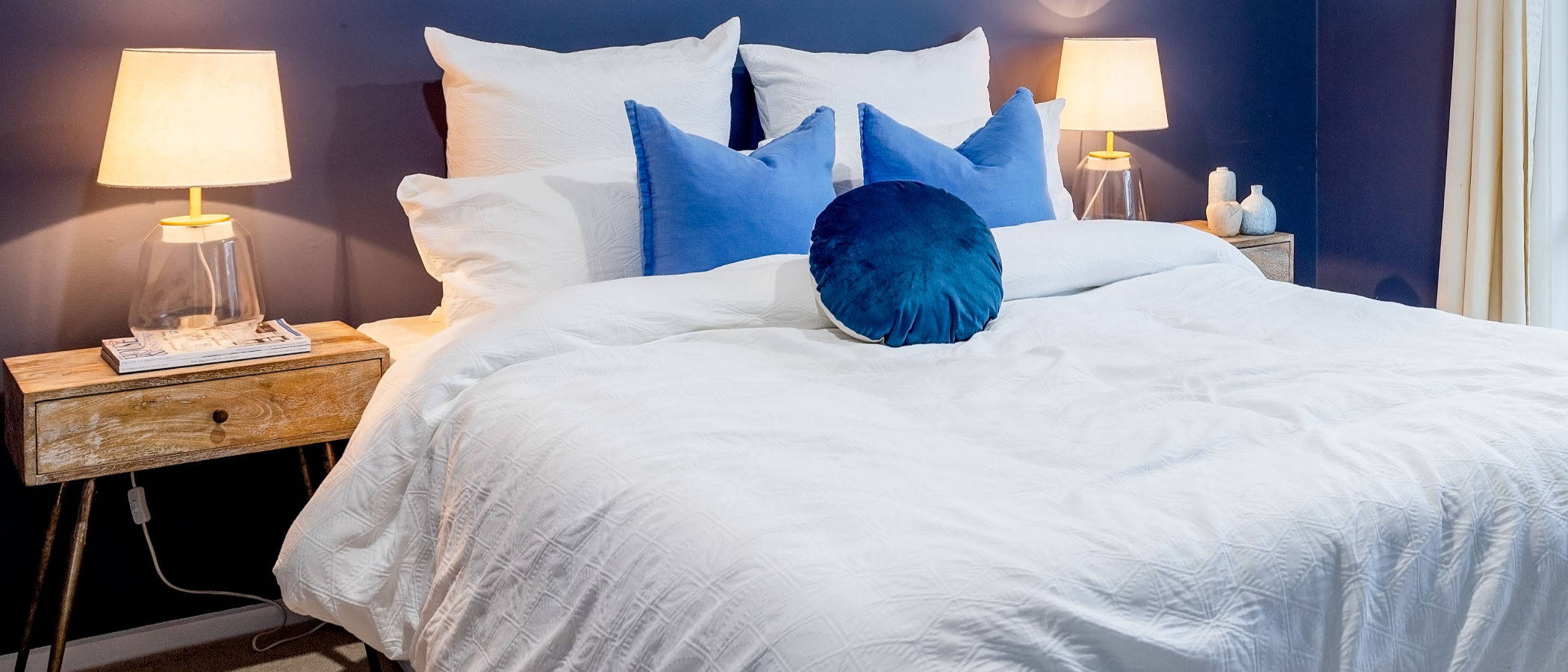A white bed with blue accent throw pillows and two lighted lamps on each side.