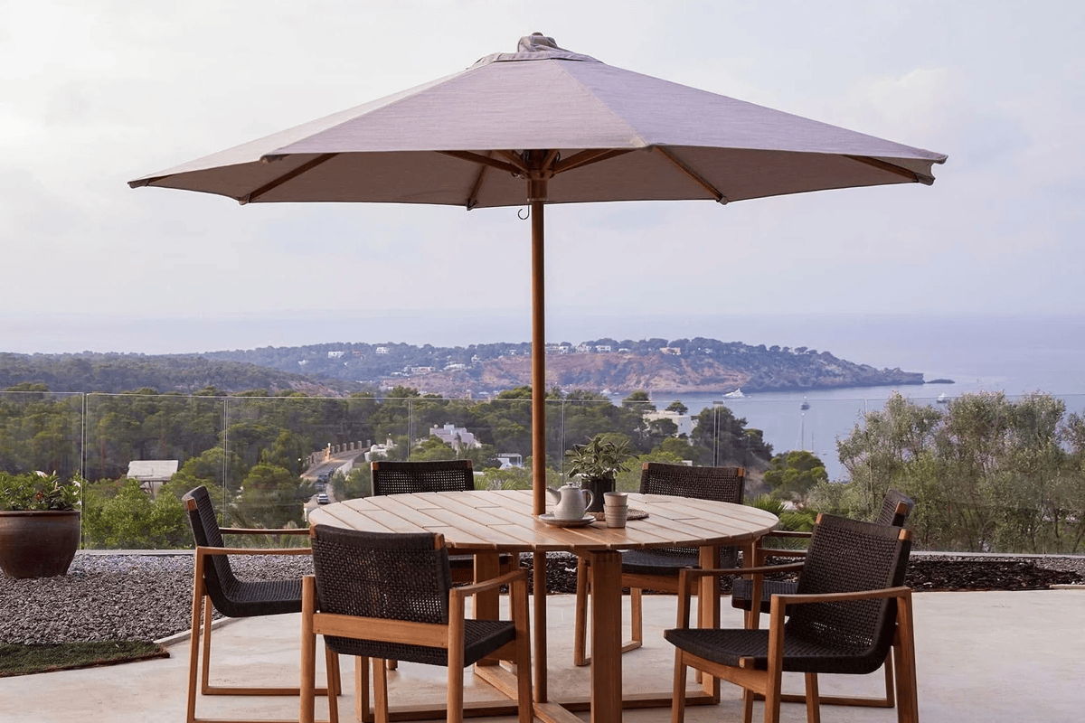 Round outdoor dining table with patio umbrella and modern dining chairs overlooking a coastal landscape.