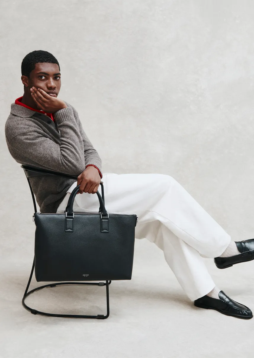 Man sitting on a chair, holding a black bag, wearing a sweater and pants.