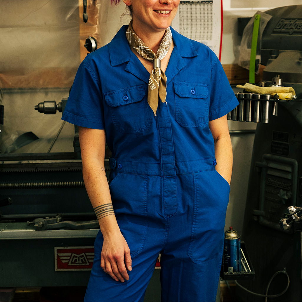 Person in blue jumpsuit smiling in a workshop setting.