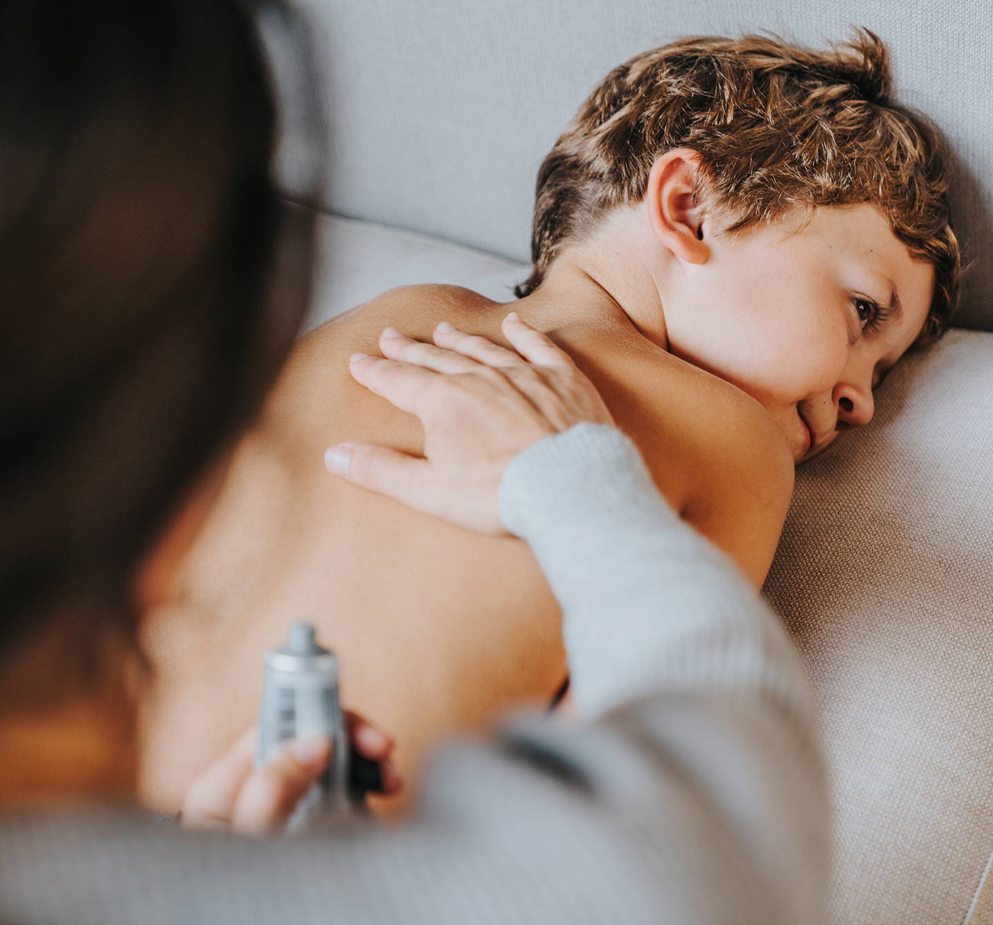Ein Junge liegt auf dem Sofa, während seine Mutter sanft ein Pflegeprodukt auf seinen Rücken aufträgt, um den Juckreiz durch Hautallergien bei Kindern zu lindern.