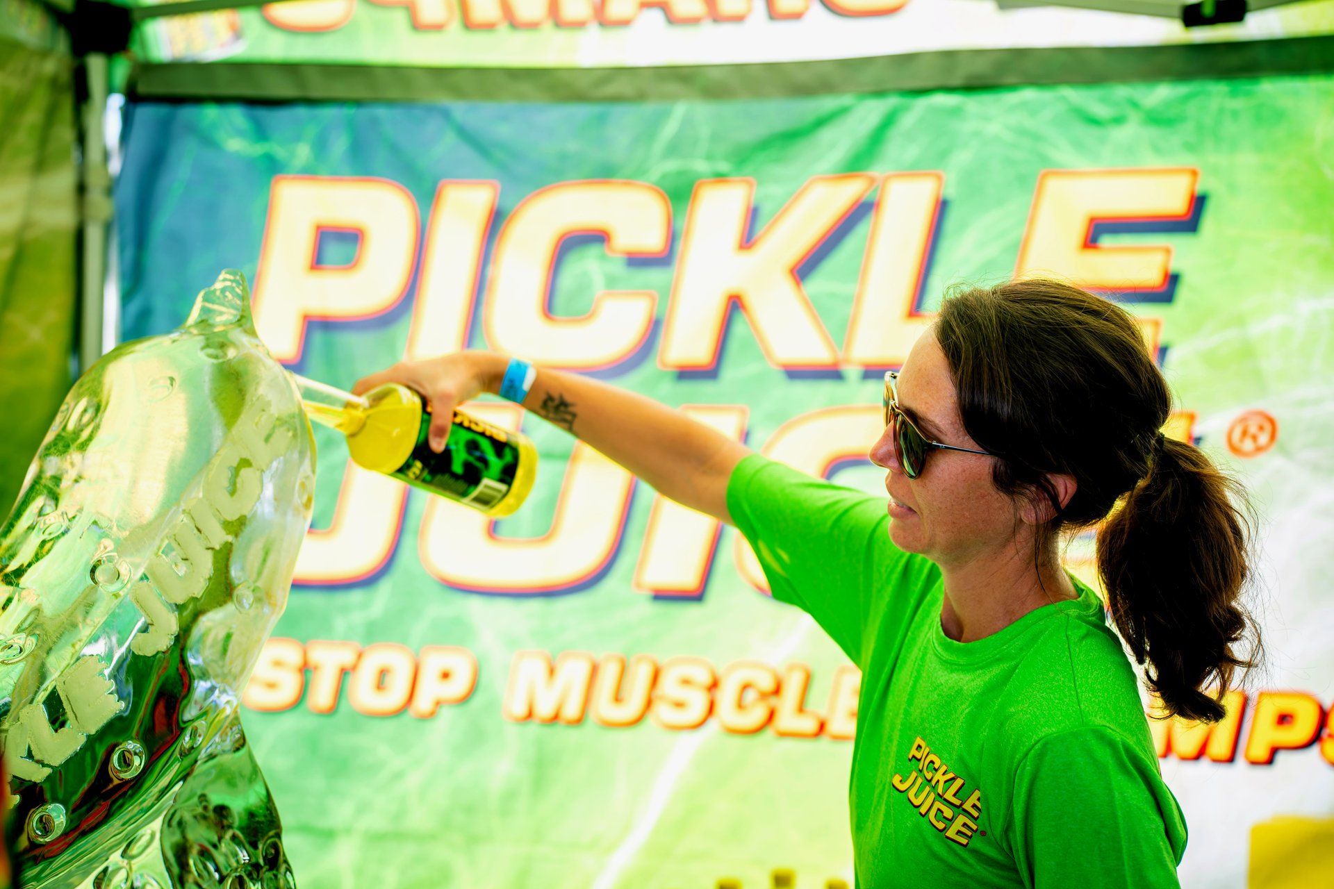 A Pickle Juice brand representative pours Pickle Juice through a custom carved pickle-shaped ice luge sculpture at the Pickle Juice 'Stop Muscle Cramps' activation at The Big Dill pickle festival.