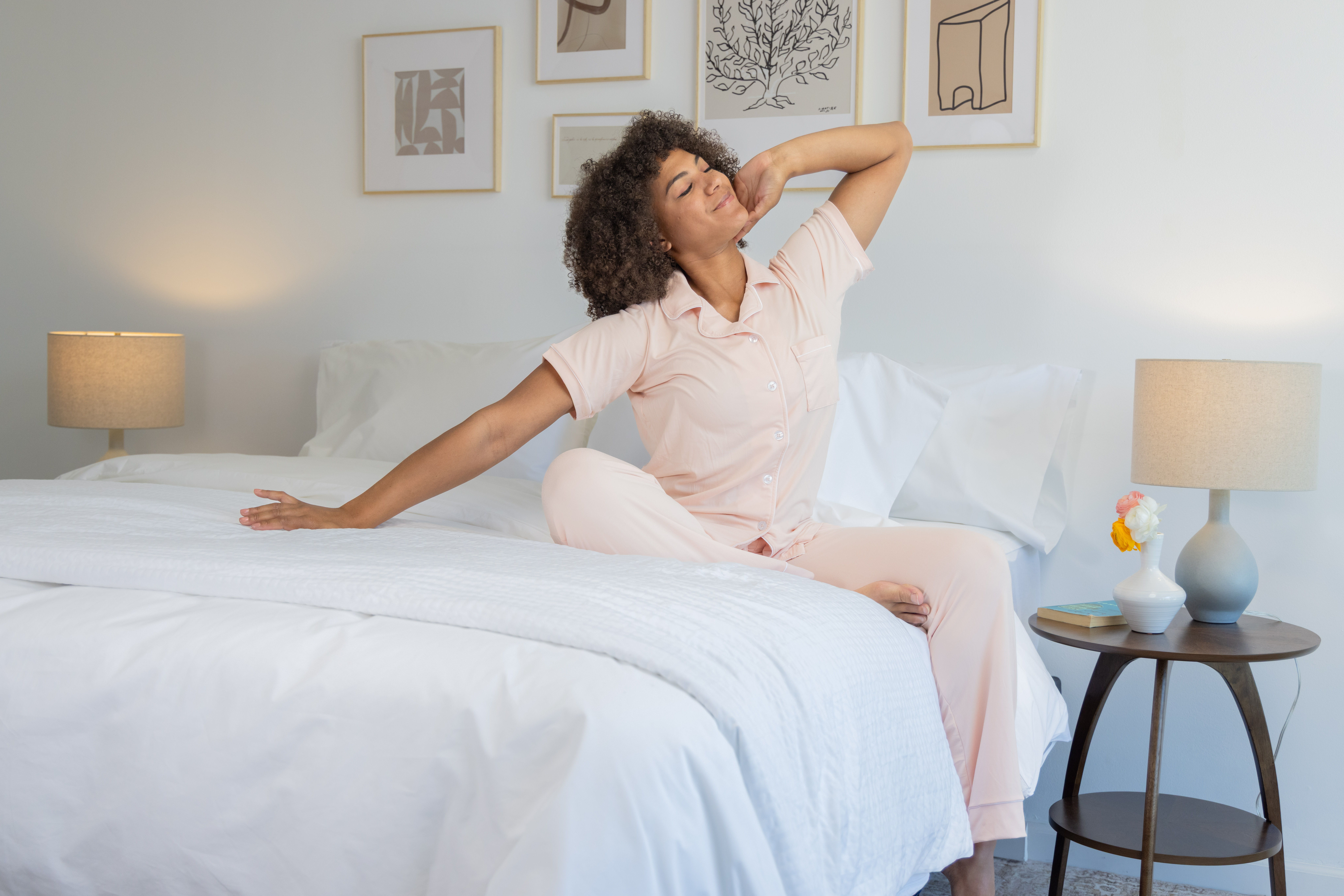 Woman stretching on the edge of her bed, white Comfortcore and Percale bedding, with wooden night tables and ceramic blue lamps