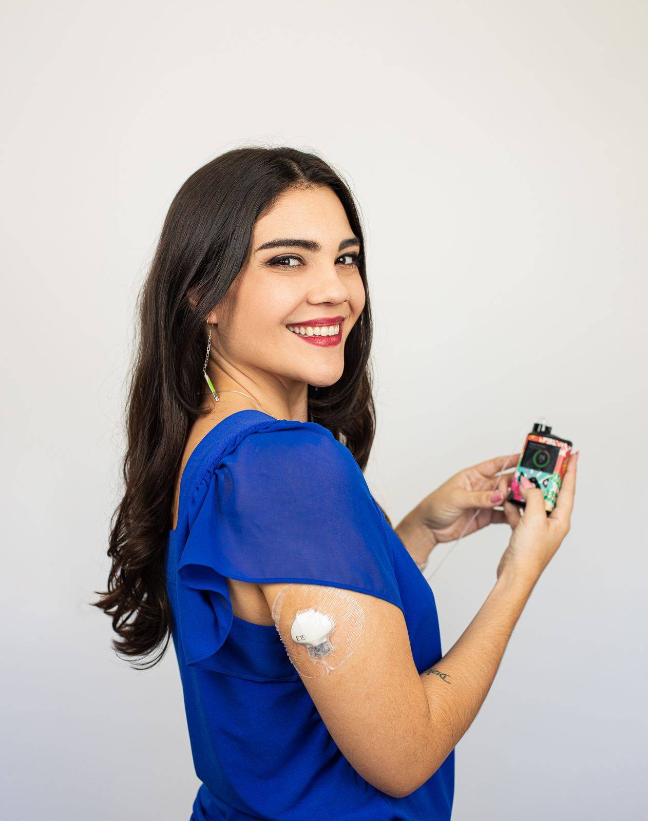 foto da Bia, uma mulher de longos cabelos pretos, com maquiagem clara e uma blusa azul royal segurando um aparelho de insulina