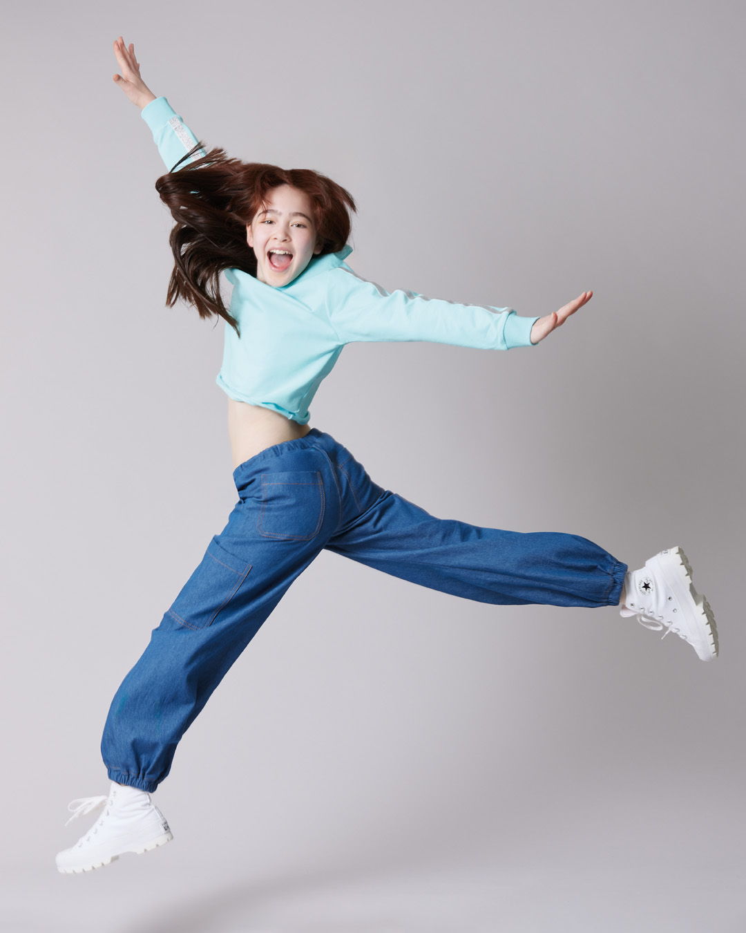 Young woman jumping joyfully in casual outfit against a gray background.