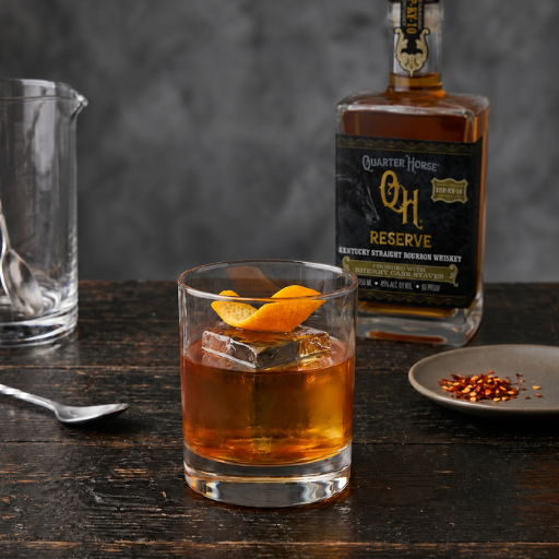 Glass of whiskey with an orange twist, bottle, and cocktail shaker on a table.